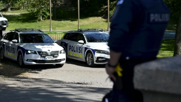 На передньому плані зображення розмитий фінський поліцейський, що спирається на стіну, а на задньому плані — ряд фінських поліцейських машин.